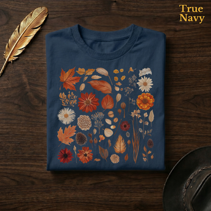 Autumn Field Guide Comfort Colors t-shirt in True Navy deep blue, folded on dark wood with gold feather quill and brown hat, displaying the full-colour botanical autumn specimen collection of russet leaves, orange dahlias, seed heads, and dried elements in warm autumn tones against the rich navy garment