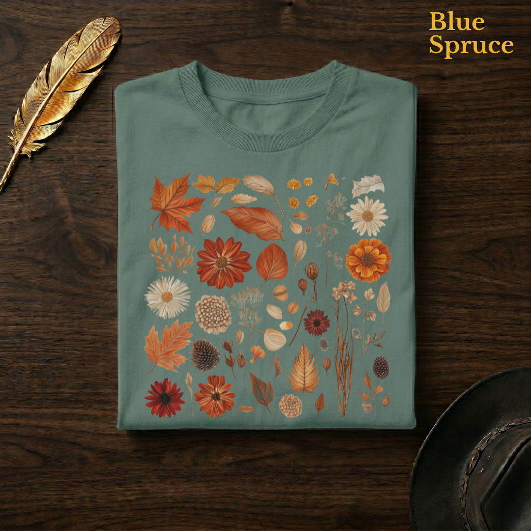 Autumn Field Guide Comfort Colors t-shirt in Blue Spruce deep teal-green, folded on dark wood with gold feather quill and brown hat, displaying the full-colour botanical autumn specimen collection of russet leaves, orange dahlias, and dried seed heads — the warm autumn palette richly contrasted against the deep teal garment — the same colourway as the park path hero shot