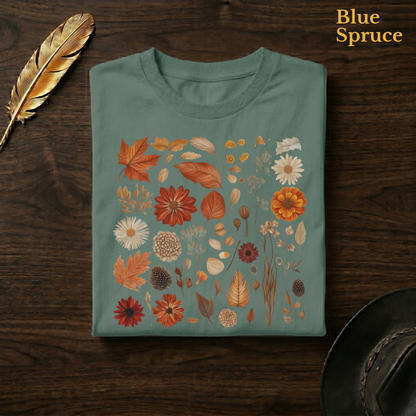 Autumn Field Guide Comfort Colors t-shirt in Blue Spruce deep teal-green, folded on dark wood with gold feather quill and brown hat, displaying the full-colour botanical autumn specimen collection of russet leaves, orange dahlias, and dried seed heads — the warm autumn palette richly contrasted against the deep teal garment — the same colourway as the park path hero shot
