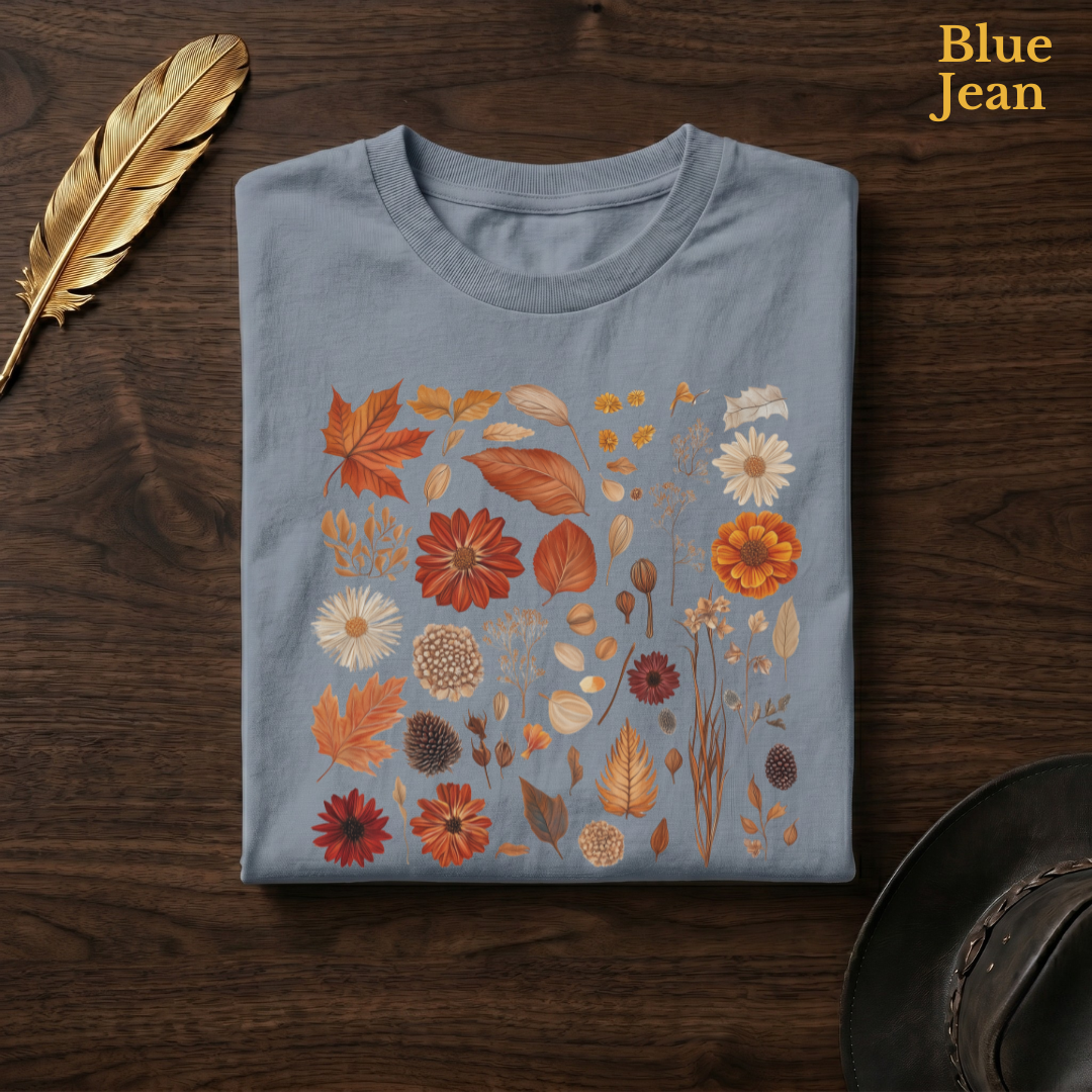 Autumn Field Guide Comfort Colors t-shirt in Blue Jean slate-grey, folded on dark wood with gold feather quill and brown hat, showing the full-colour watercolour botanical autumn specimen collection of leaves, flowers, pine cones, and seed heads against the washed slate garment — the same colourway as the cafe hero shot