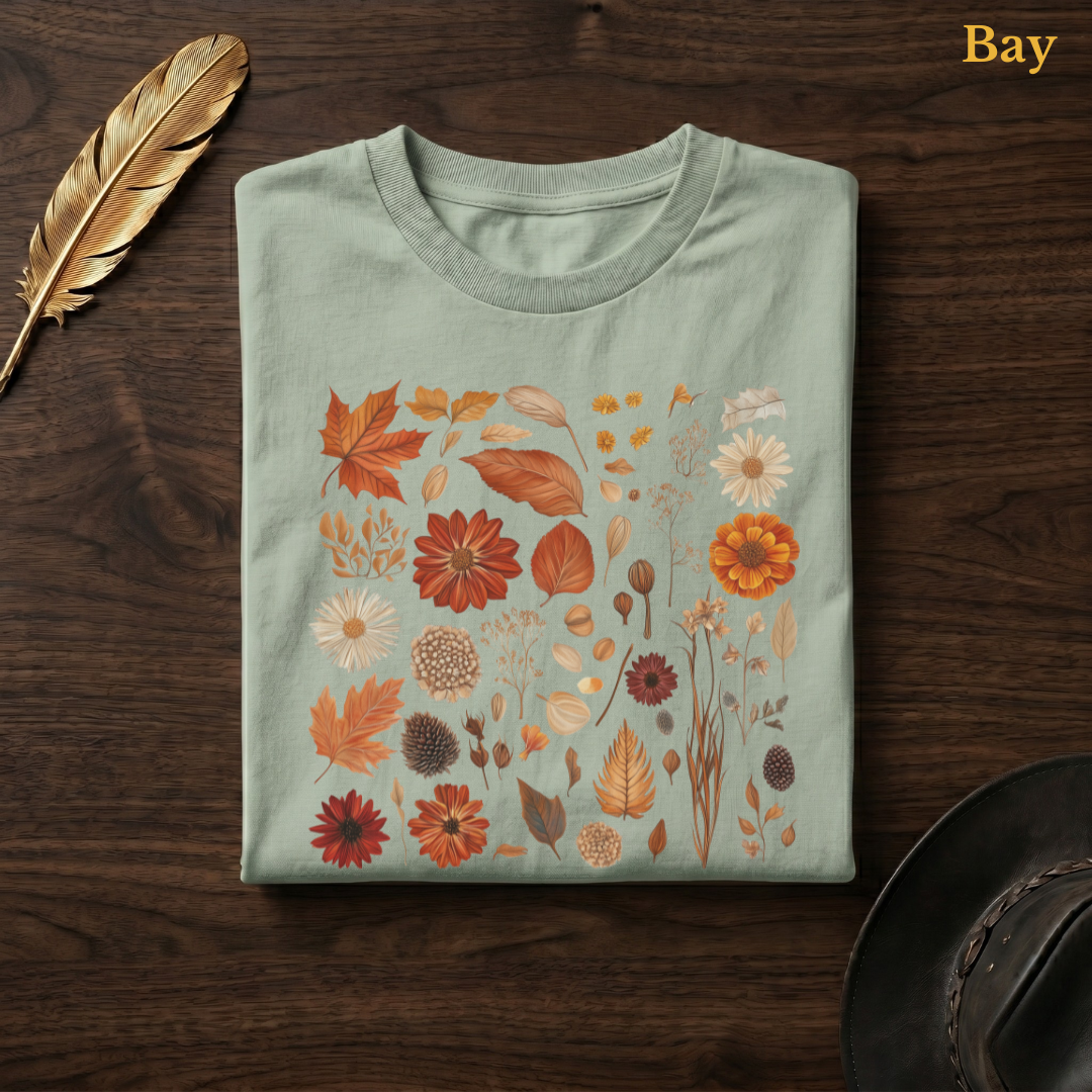 Autumn Field Guide Comfort Colors garment-dyed t-shirt in Bay sage-green, folded on dark wood with gold feather quill and brown hat, showing the full-colour watercolour botanical specimen collection of autumn leaves, dahlias, chrysanthemums, pine cones, seed heads, and dried elements in warm terracotta and russet tones