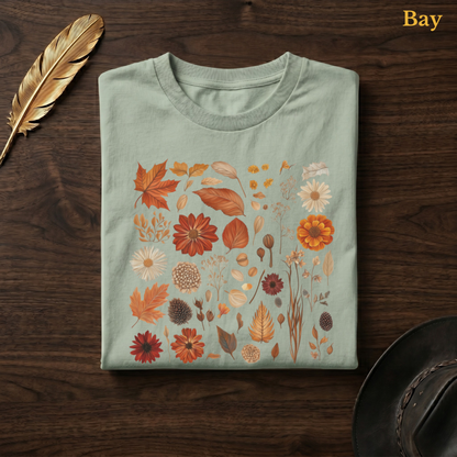Autumn Field Guide Comfort Colors garment-dyed t-shirt in Bay sage-green, folded on dark wood with gold feather quill and brown hat, showing the full-colour watercolour botanical specimen collection of autumn leaves, dahlias, chrysanthemums, pine cones, seed heads, and dried elements in warm terracotta and russet tones