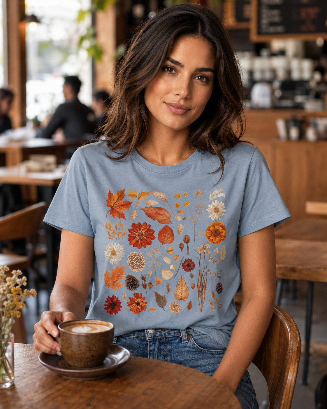 Smiling woman wearing the Autumn Field Guide Comfort Colors t-shirt in Blue Jean slate-grey, sitting at a cafe table with a latte in a warm wooden cafe interior with a chalkboard menu visible behind her — the full-colour botanical autumn specimen collection of leaves, flowers, and seed heads showing warmly against the slate garment in a cosy seasonal setting