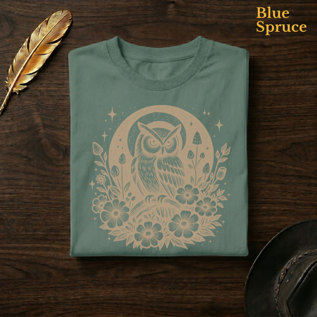 Celestial Owl Full Moon Comfort Colors t-shirt in Blue Spruce deep teal-green, folded on dark wood with gold feather quill and brown hat, displaying the warm amber celestial owl and full moon wildflower folk art illustration against the rich deep garment