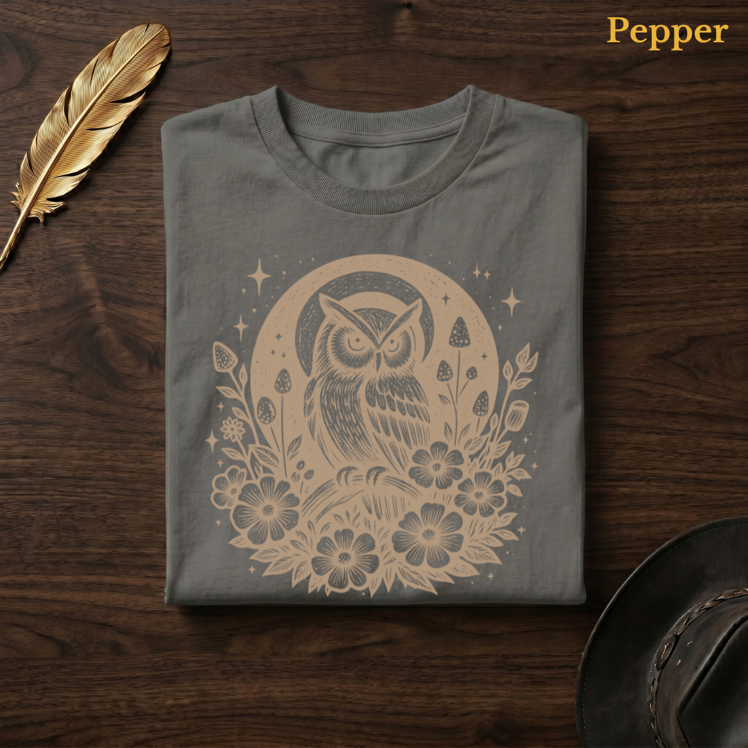 Celestial Owl Full Moon Comfort Colors t-shirt in Pepper olive-charcoal, folded on dark wood with gold feather quill and brown hat, showing the warm amber celestial owl and full moon wildflower folk art design against the deep earthy garment