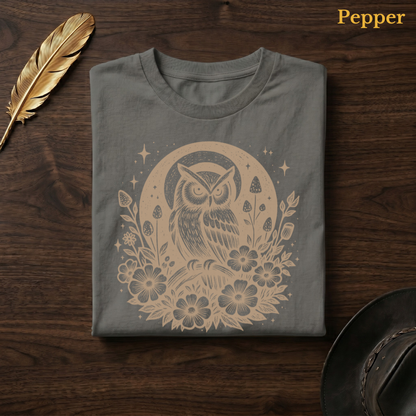 Celestial Owl Full Moon Comfort Colors t-shirt in Pepper olive-charcoal, folded on dark wood with gold feather quill and brown hat, showing the warm amber celestial owl and full moon wildflower folk art design against the deep earthy garment