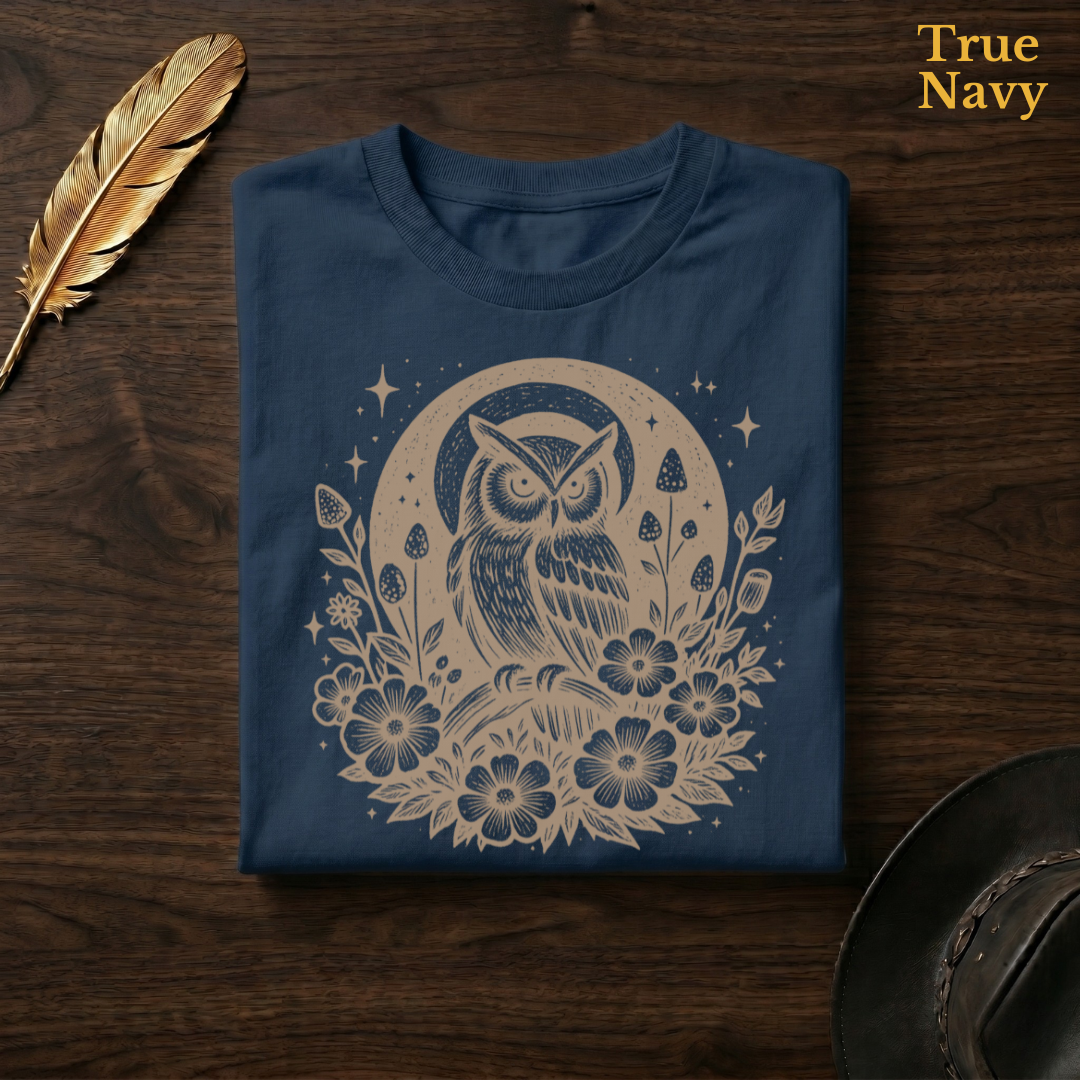 Celestial Owl Full Moon Comfort Colors t-shirt in True Navy deep blue, folded on dark wood with gold feather quill and brown hat, displaying the warm amber celestial owl and full moon wildflower folk art illustration against the rich navy garment