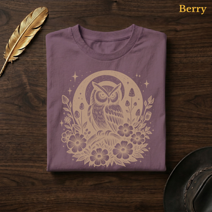 Celestial Owl Full Moon Comfort Colors t-shirt in Berry mauve, folded on dark wood with gold feather quill and brown hat, displaying the warm earthy celestial folk art owl and full moon wildflower illustration — the same colourway as the sofa hero shot
