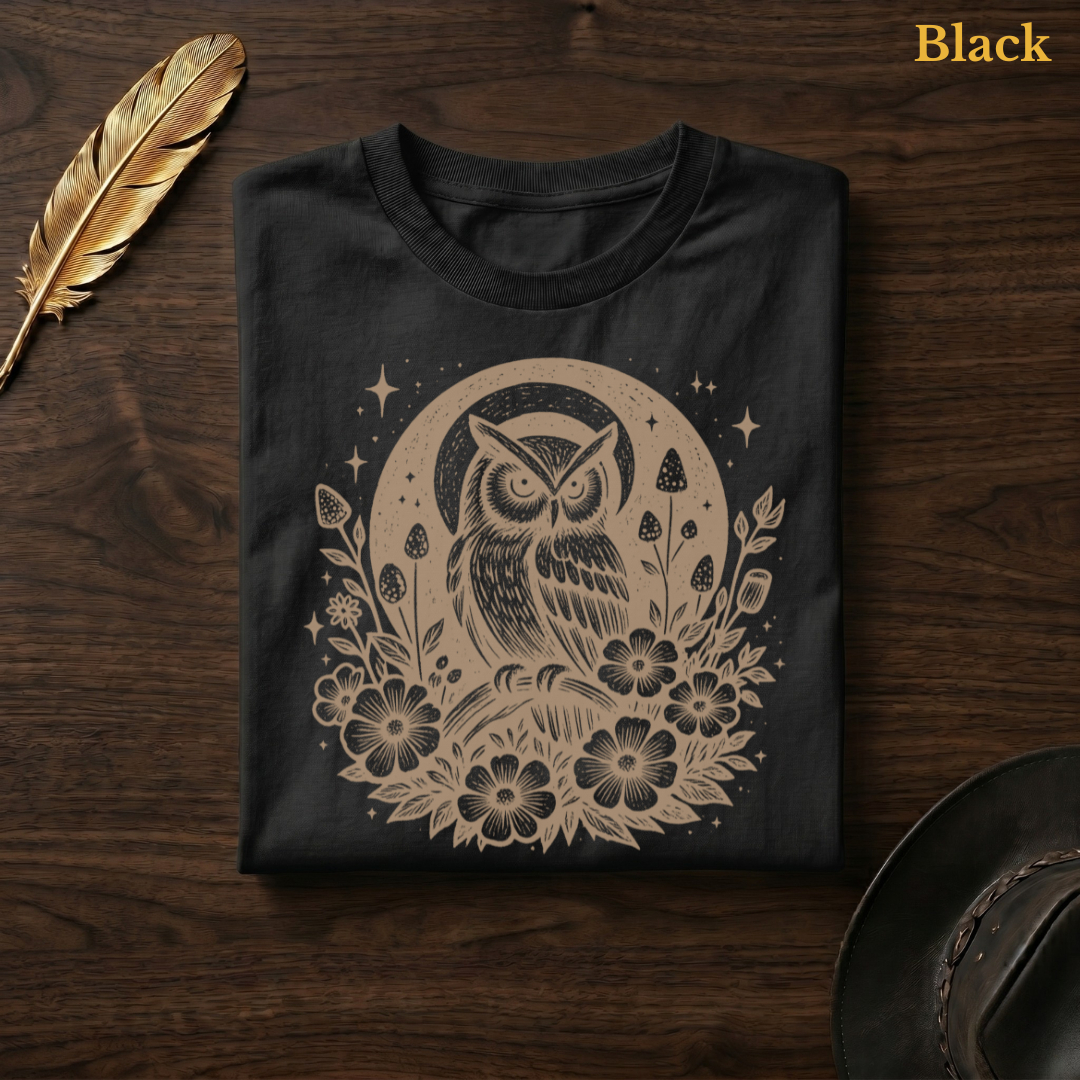 Celestial Owl Full Moon Comfort Colors t-shirt in Black, folded on dark wood with gold feather quill and brown hat, showing the celestial owl and full moon wildflower folk art design in deep amber tone — the sparkle stars and open blooms glowing warmly against the dark garment