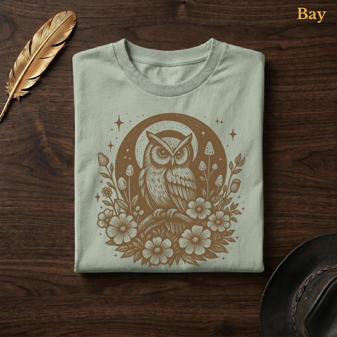 Celestial Owl Full Moon Comfort Colors garment-dyed t-shirt in Bay sage-green, folded on dark wood with gold feather quill and brown hat, showing the folk art illustration of an owl at the centre of a glowing full moon with sparkle stars and large open wildflowers