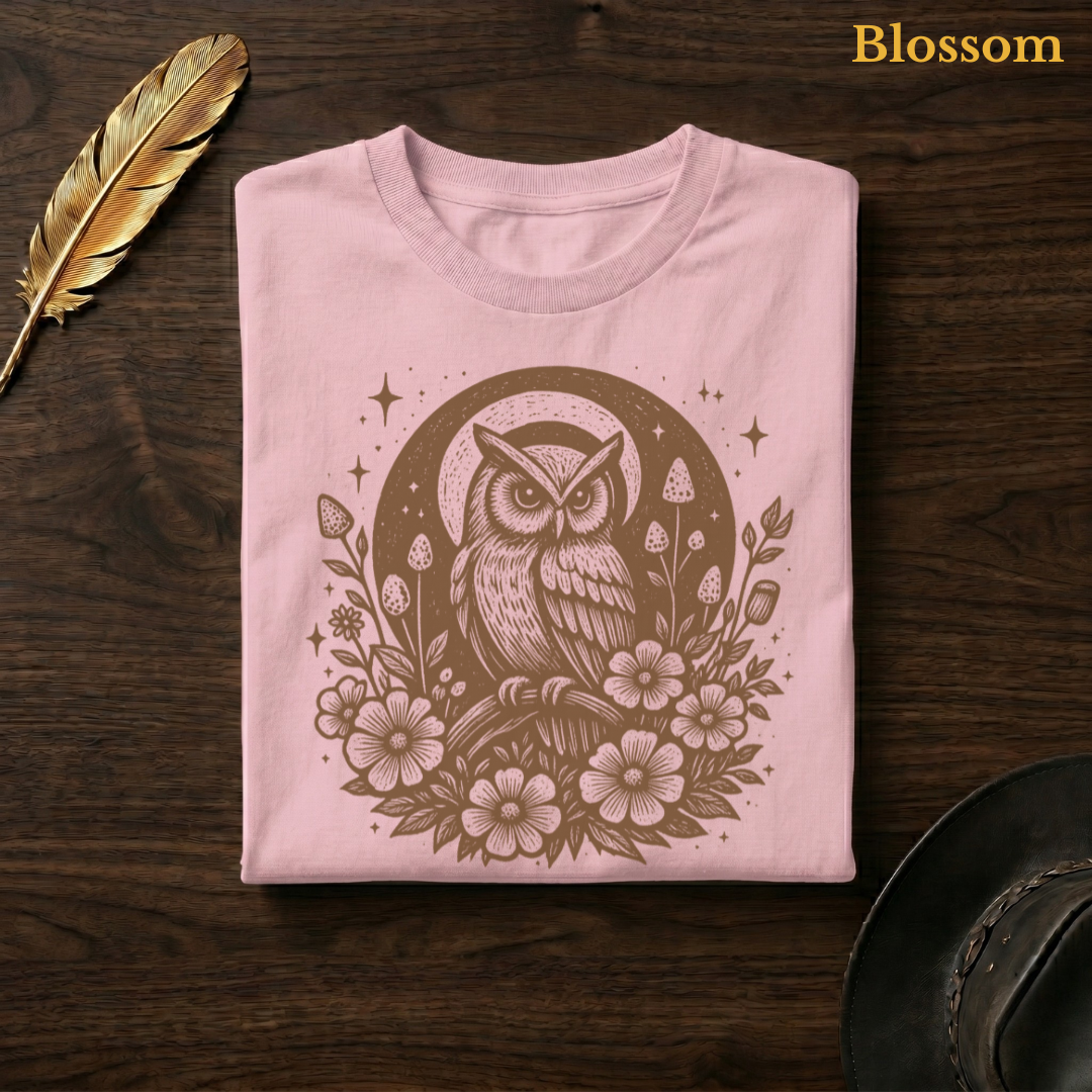 Celestial Owl Full Moon Comfort Colors garment-dyed tee in Blossom soft pink, folded on dark wood with gold feather quill and brown hat, displaying the warm-toned celestial owl and full moon wildflower folk art illustration on a delicate pink garment — the same colourway as the window sofa hero shot