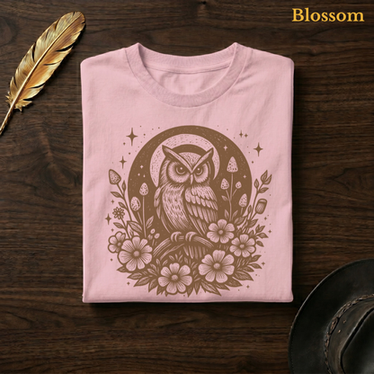 Celestial Owl Full Moon Comfort Colors garment-dyed tee in Blossom soft pink, folded on dark wood with gold feather quill and brown hat, displaying the warm-toned celestial owl and full moon wildflower folk art illustration on a delicate pink garment — the same colourway as the window sofa hero shot
