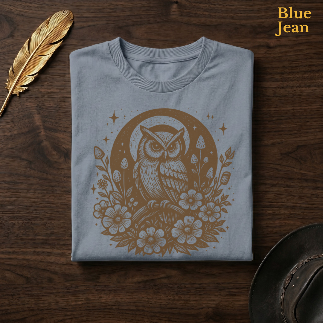 Celestial Owl Full Moon Comfort Colors t-shirt in Blue Jean slate-grey, folded on dark wood with gold feather quill and brown hat, showing the celestial owl at the heart of a full moon with sparkle stars and wildflowers in warm earthy tone