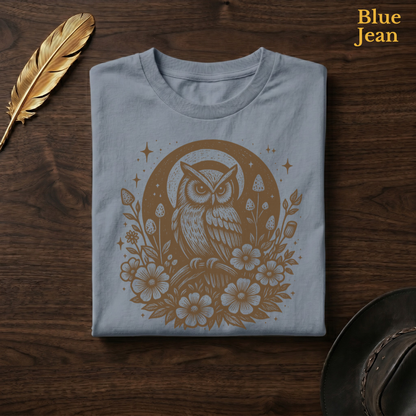 Celestial Owl Full Moon Comfort Colors t-shirt in Blue Jean slate-grey, folded on dark wood with gold feather quill and brown hat, showing the celestial owl at the heart of a full moon with sparkle stars and wildflowers in warm earthy tone