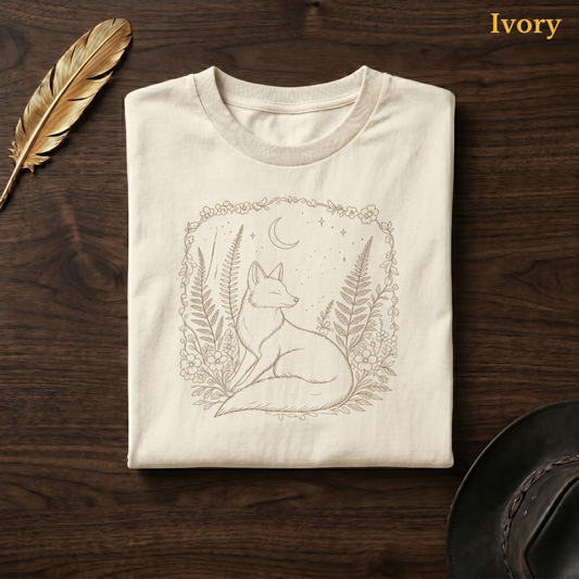Boho Fox & Forest Flowers Comfort Colors t-shirt in Ivory cream, flat lay on dark wood with gold feather quill, displaying the warm sandy fox and forest folk art illustration on a pale cream garment — the same colourway as the hydrangea garden and boho bench hero shots