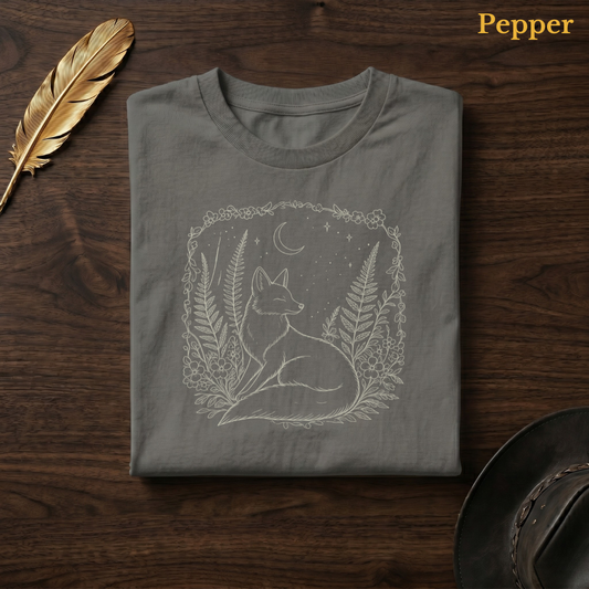 Boho Fox & Forest Flowers Comfort Colors t-shirt in Pepper olive-charcoal, flat lay on dark wood with gold feather quill, displaying the warm amber fox and forest folk art illustration — the same colourway as the rose garden hero shot