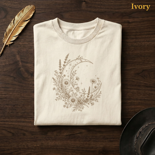 Moon & Wildflowers Comfort Colors t-shirt in Ivory cream, flat lay on dark wood with gold feather quill, displaying the warm sandy botanical moon and wildflower illustration on a pale cream garment — the same colourway as the gardening hero shot