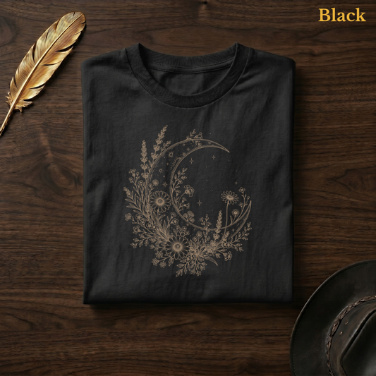 Moon & Wildflowers Comfort Colors t-shirt in Black, flat lay on dark wood with gold feather quill, showing the wildflower moon botanical illustration in deep amber tone against the dark garment — the same colourway as the wildflower field hero shot