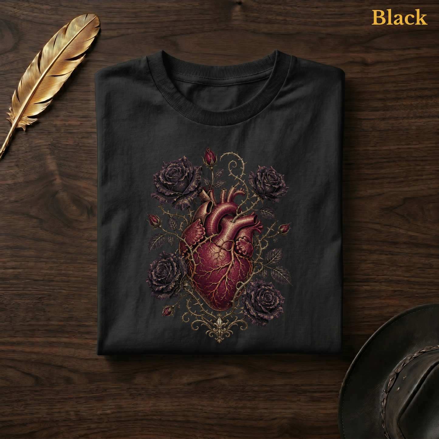 Anatomical Heart in Bloom tee in Black, folded flat on dark walnut wood beside a gold feather and a leather hat, the anatomical heart, thorn vine, and rose design printed on the chest, labelled Black.