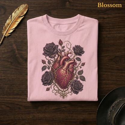 Anatomical Heart in Bloom tee in Blossom, a soft pastel pink garment-dyed shirt folded flat on dark walnut wood beside a gold feather and a leather hat, the gothic botanical heart design on the chest, labelled Blossom.