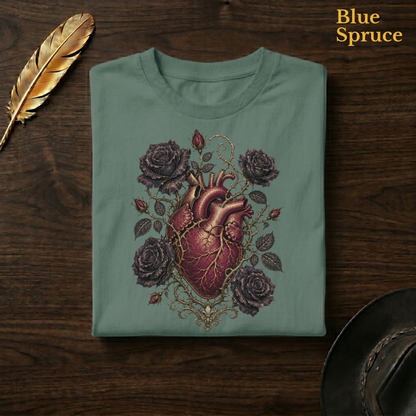 Anatomical Heart in Bloom tee in Blue Spruce, a deep teal-green garment-dyed shirt folded flat on dark walnut wood beside a gold feather and a leather hat, the morbid anatomy heart design on the chest, labelled Blue Spruce.