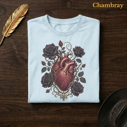 Anatomical Heart in Bloom tee in Chambray, a soft pale blue garment-dyed shirt folded flat on dark walnut wood beside a gold feather and a leather hat, the anatomical heart and rose design on the chest, labelled Chambray.