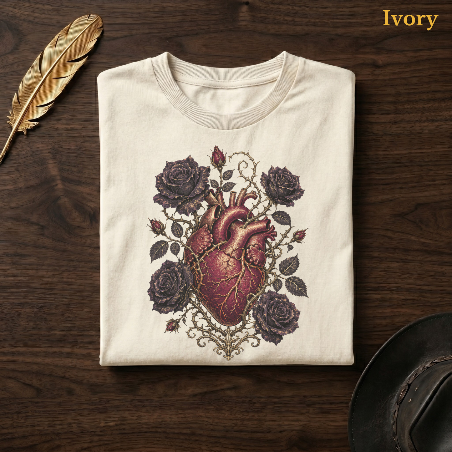 Anatomical Heart in Bloom tee in Ivory, folded flat on dark walnut wood beside a gold feather and a leather hat, the gothic heart, thorn vine, and black rose design on the chest, labelled Ivory.