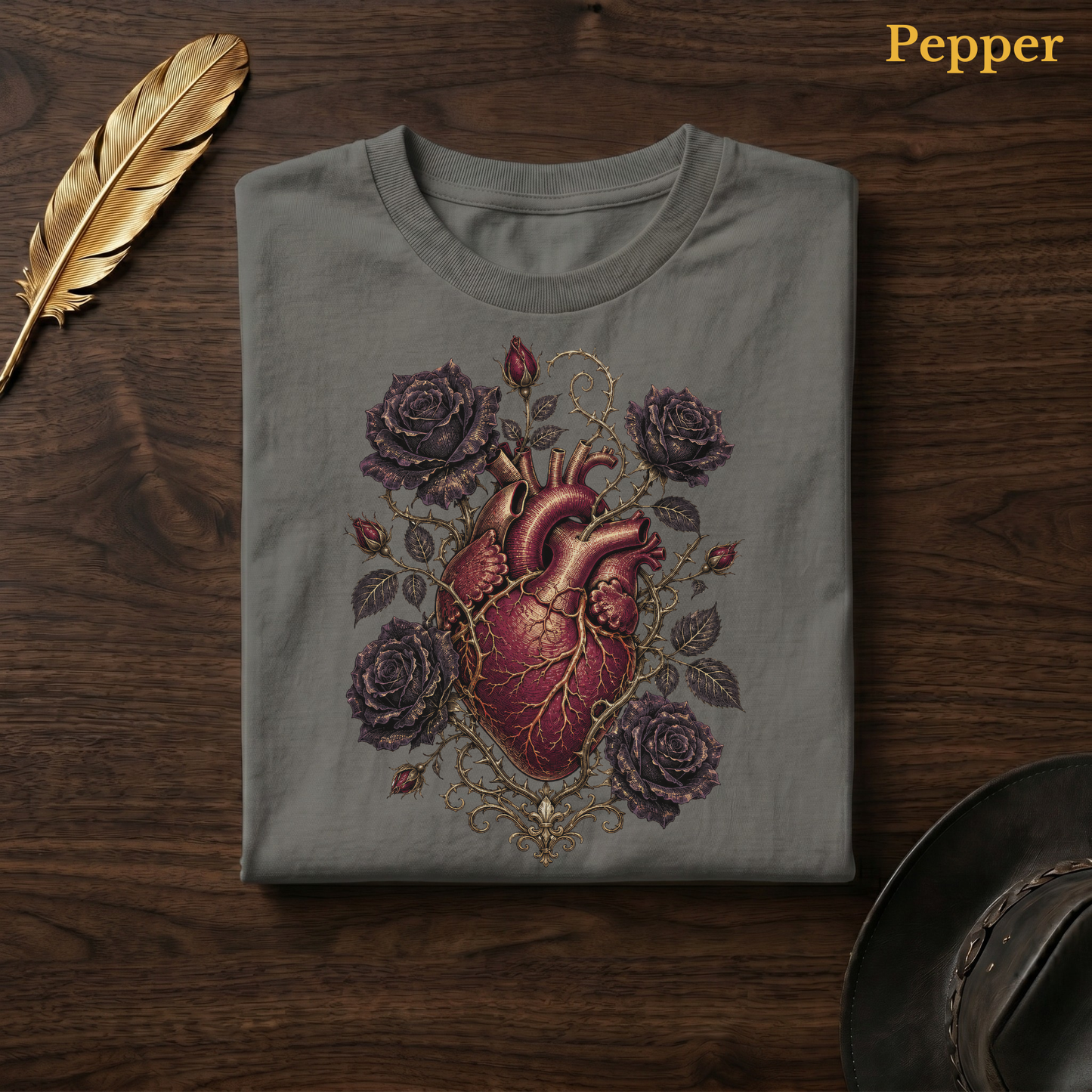 Anatomical Heart in Bloom tee in Pepper, a warm grey-charcoal garment-dyed shirt folded flat on dark walnut wood beside a gold feather and a leather hat, the anatomical heart design centred on the chest, labelled Pepper.
