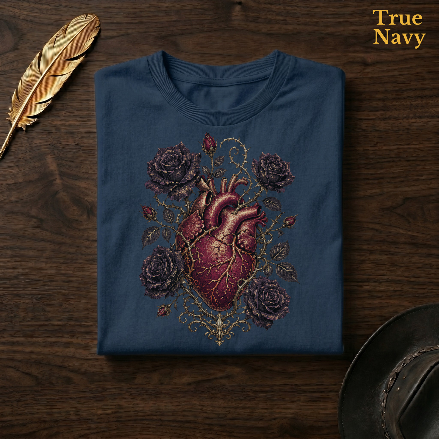 Anatomical Heart in Bloom tee in True Navy, a deep navy garment-dyed shirt folded flat on dark walnut wood beside a gold feather and a leather hat, the gothic botanical heart design on the chest, labelled True Navy.