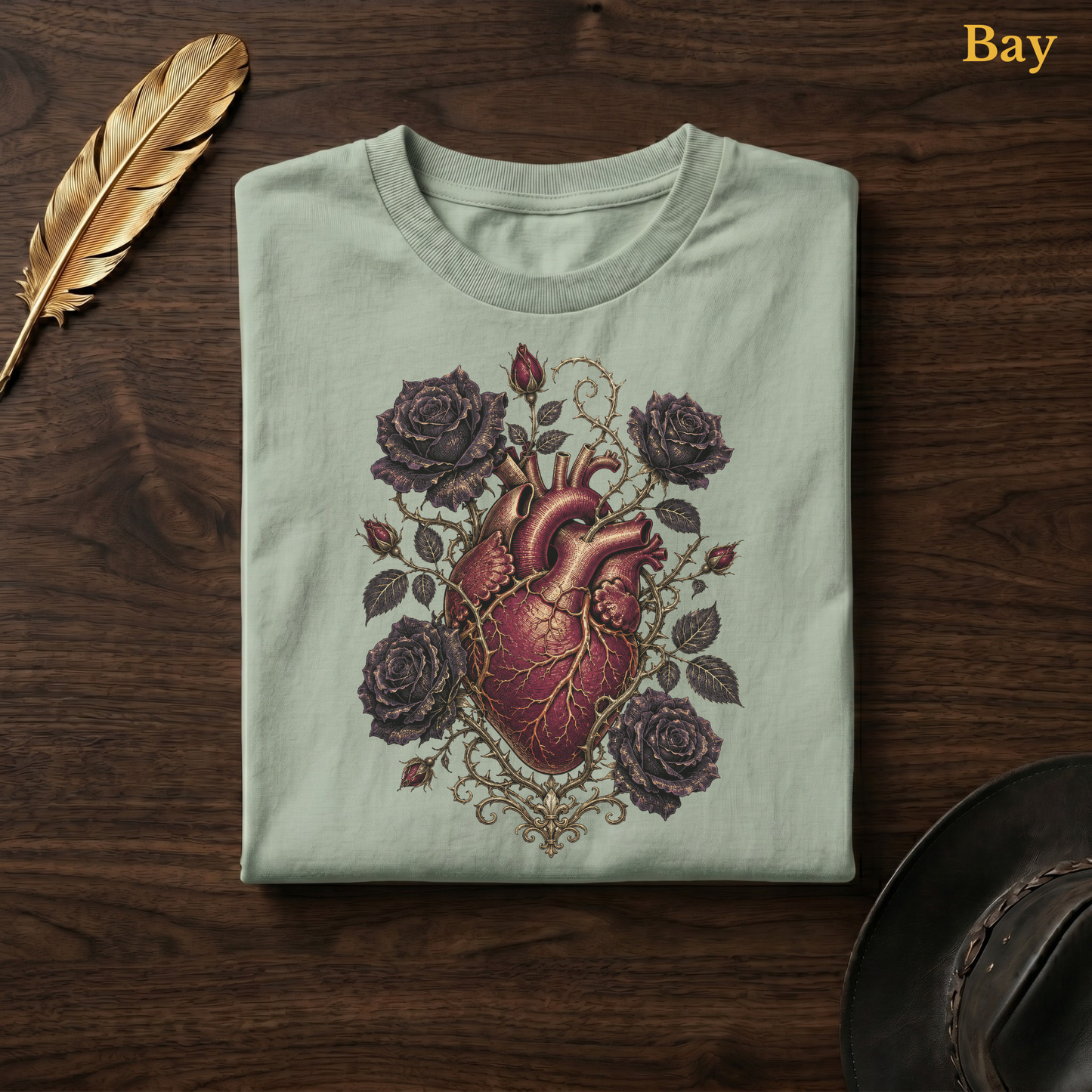 Anatomical Heart in Bloom tee in Bay, a soft sage-green garment-dyed shirt folded flat on dark walnut wood beside a gold feather and a leather hat, the gothic heart and rose design printed on the chest, labelled Bay.