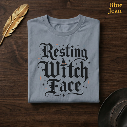 Resting Witch Face tee in Blue Jean, a faded denim-blue garment-dyed shirt folded flat on dark walnut wood beside a gold feather and a leather hat, the sassy halloween lettering design on the chest, labelled Blue Jean.