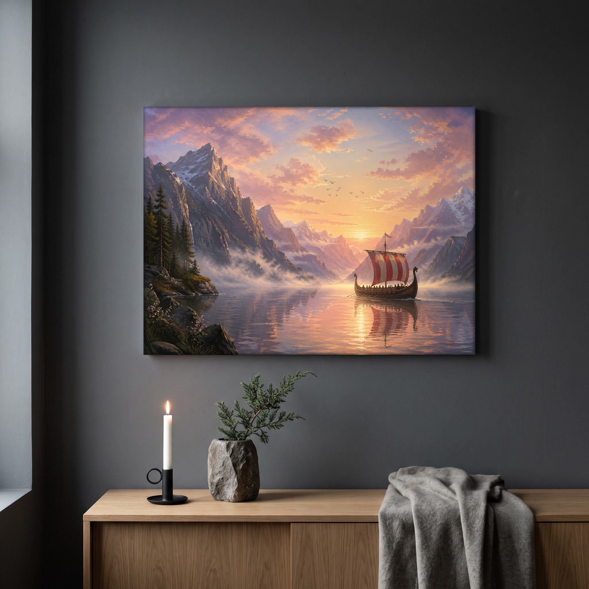 The Longship at Dawn fjord print hanging on a plain dark grey wall above a light oak sideboard, styled with a lit white taper candle in a black holder, a stone vase of juniper, and a soft grey throw draped over the end.