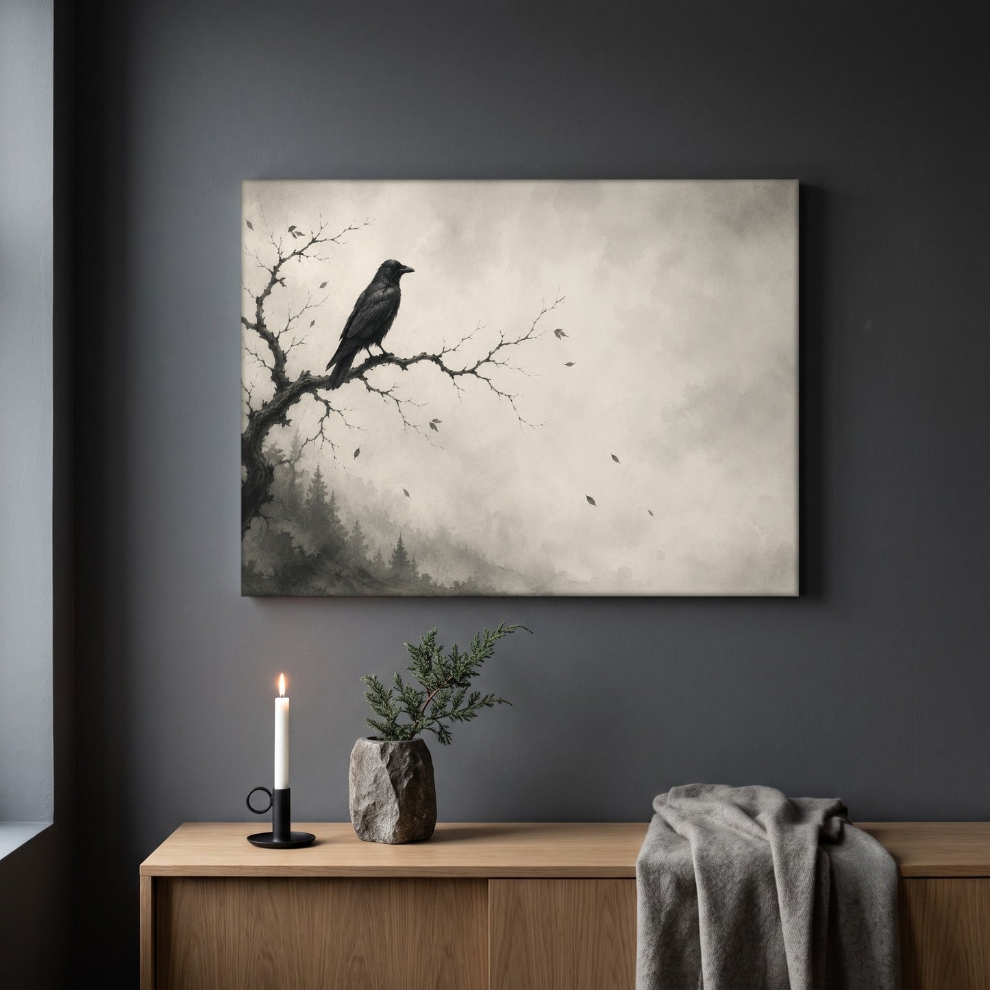 Hollow Morning raven print hanging on a plain dark grey wall above a light oak sideboard, styled with a lit white taper candle in a black holder, a stone vase of juniper, and a soft grey throw draped over the end.