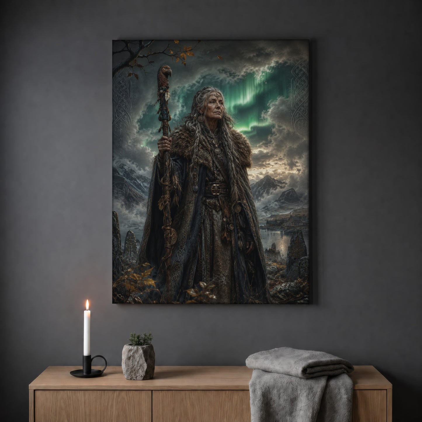 Spákona seeress print hanging on a plain grey wall above a light oak sideboard, styled with a lit white taper candle in a black holder, a small stone pot of greenery and a folded grey throw.