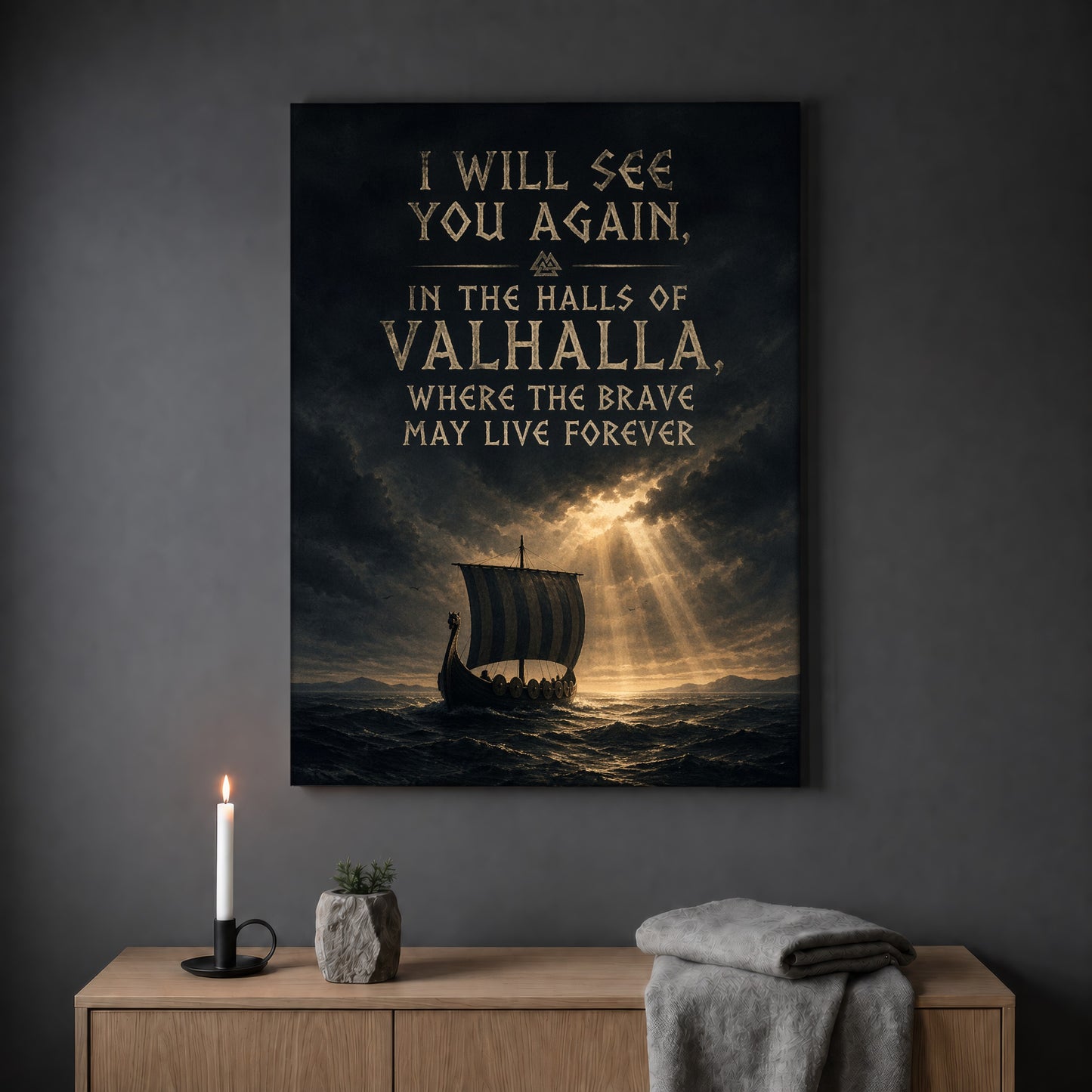 Valhalla Awaits quote print hanging on a plain grey wall above a light oak sideboard, styled with a lit white taper candle in a black holder, a small stone pot of greenery and a folded grey throw.