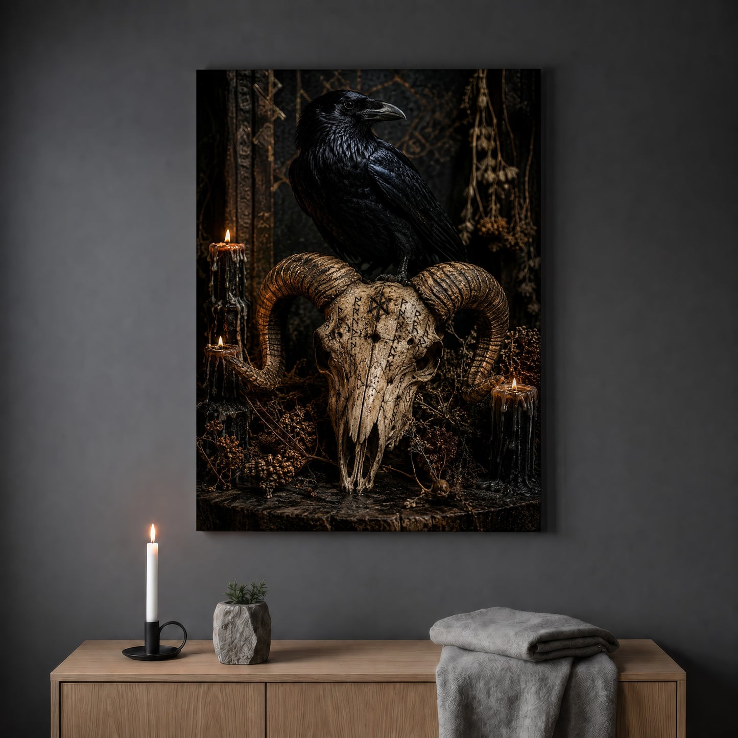 Valravn raven skull print hanging on a plain grey wall above a light oak sideboard, styled with a lit white taper candle in a black holder, a small stone pot of greenery and a folded grey throw.
