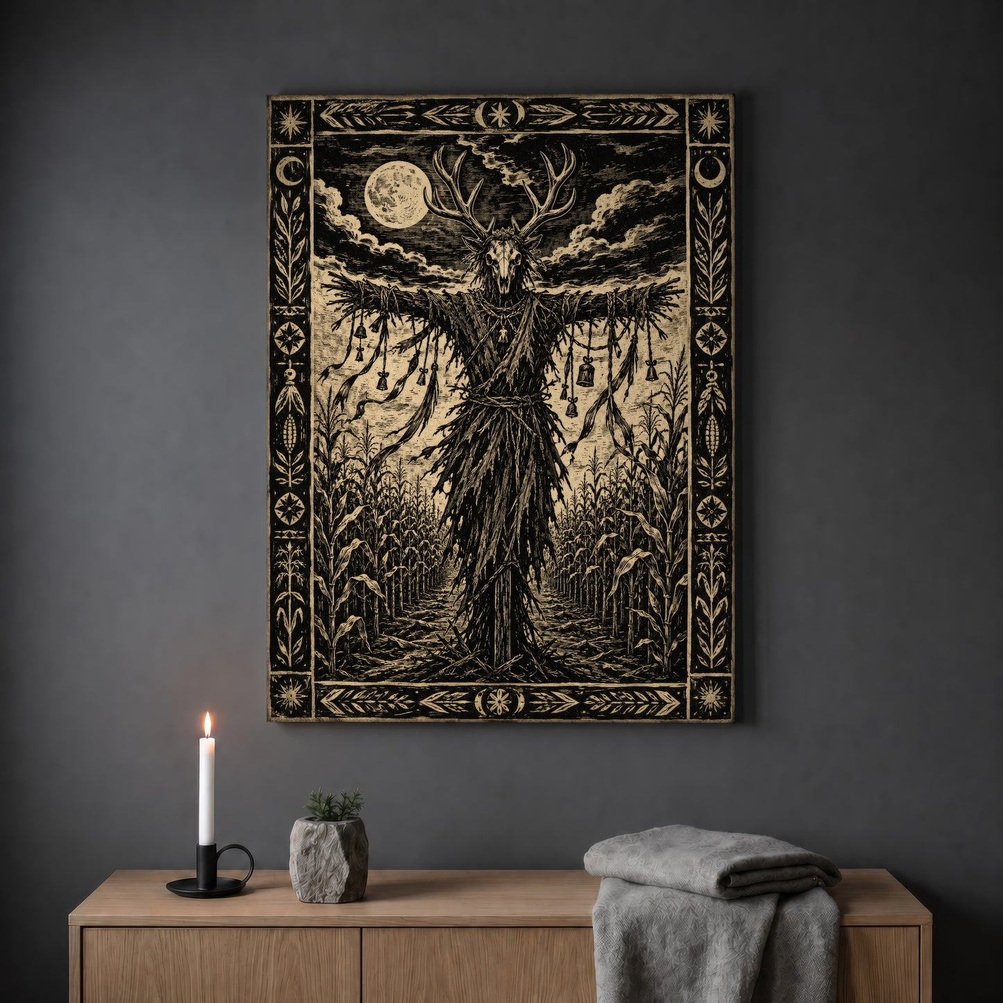 The Corn Sentinel woodcut print hanging on a plain grey wall above a light oak sideboard, styled with a lit white taper candle in a black holder, a small stone pot of greenery and a folded grey throw.