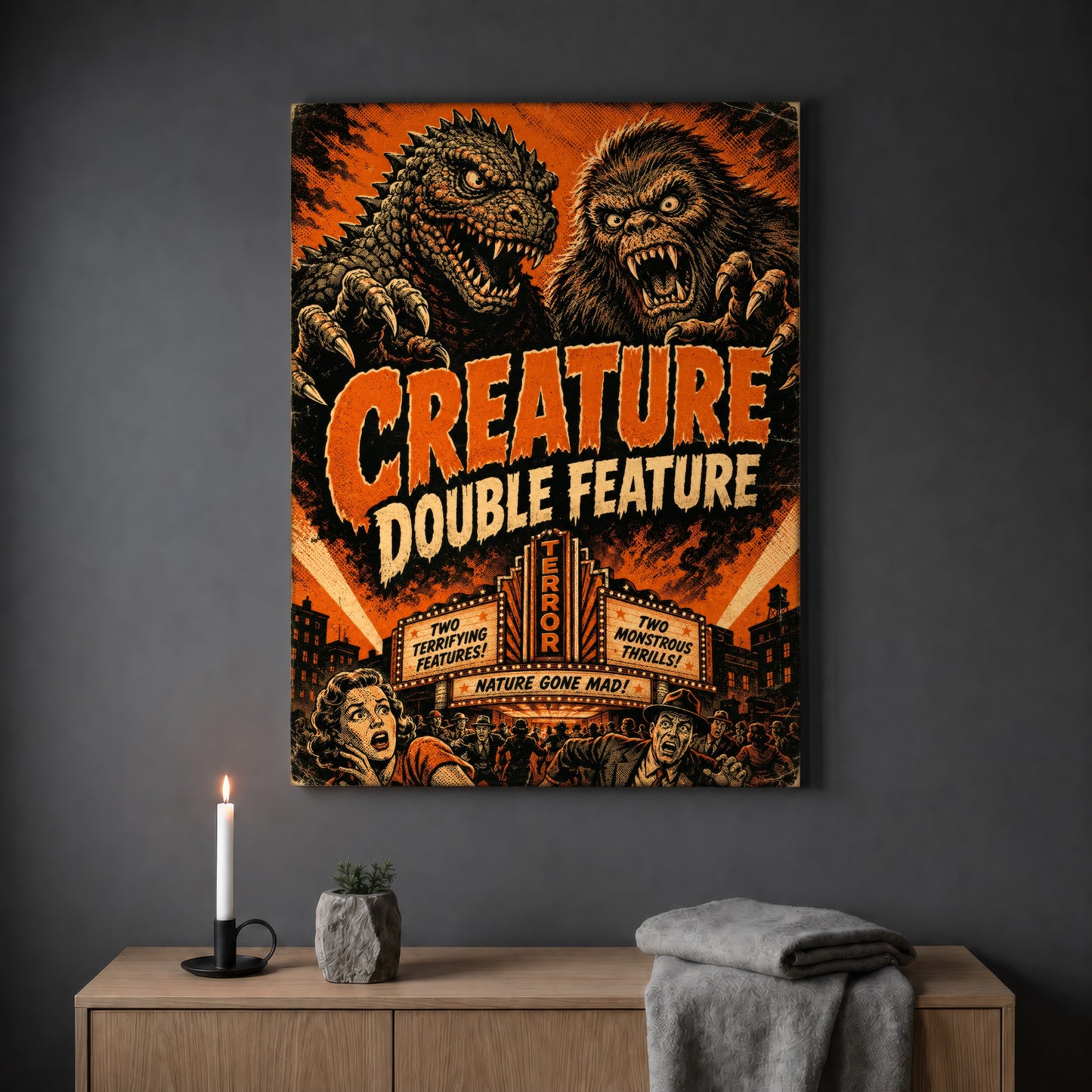 Marquee Mayhem kaiju poster hanging on a plain grey wall above a light oak sideboard, styled with a lit white taper candle in a black holder, a small stone pot of greenery and a folded grey throw.